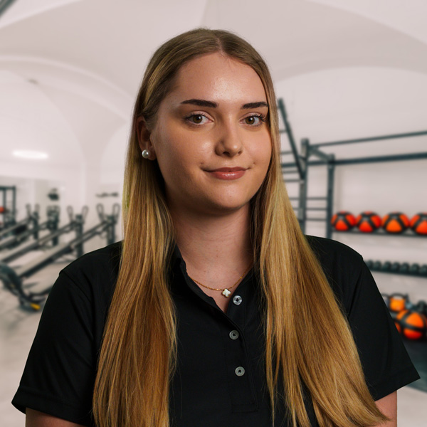 Jessica Ma mit langen blonden Haaren, trägt ein schwarzes Polohemd, vor Trainingsgeräten in einer hellen Therapieräumung der f+p Gesund Bewegen im Allgäu