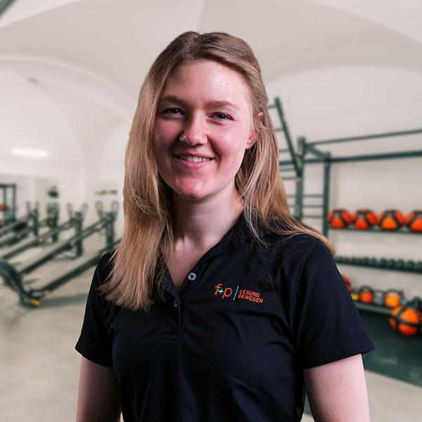 Josefine, Mitarbeiterin von f+p Gesund Bewegen im Allgäu, lächelt freundlich. Sie trägt ein dunkles Polo mit dem Logo von f+p; im Hintergrund befinden sich Fitnessgeräte wie Hanteln und Medizinbälle in einem hellen Trainingsraum.