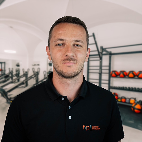 Josip, Mitarbeitender von f+p Gesund Bewegen, im Allgäu-Praxen Studio. Porträt eines Mannes mit schwarzem Polo vor Trainingsgeräten, zentrale Blickführung, helle Räumlichkeiten.