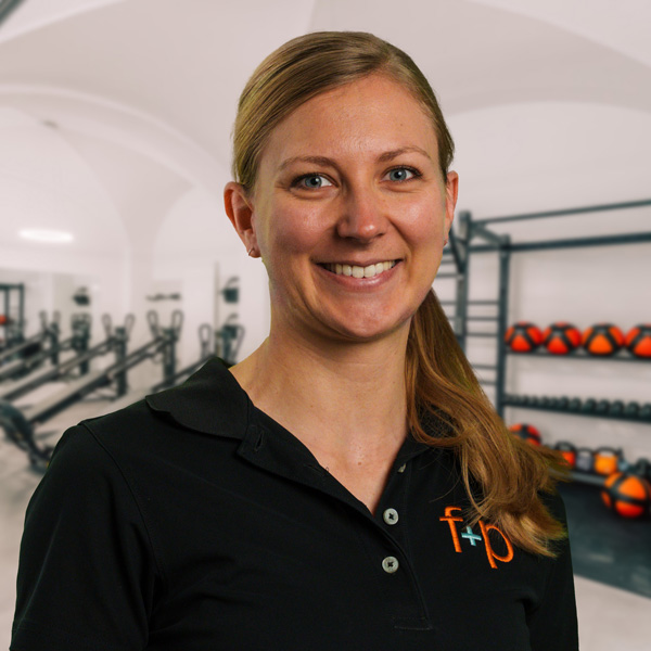 Katrin, blonde Physiotherapeutin in schwarzer Polo-Bluse, lächelt freundlich in einem Fitness-Studio mit Geräten und Reha-Ausstattung im Hintergrund