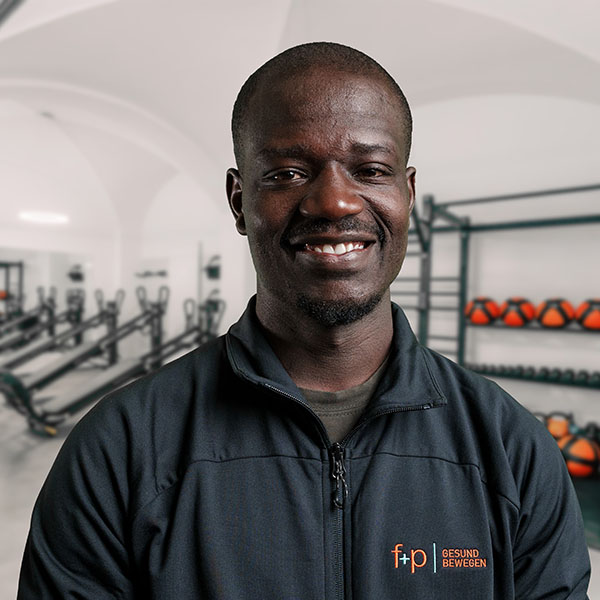 LucasOc Mann in schwarzer Sportjacke mit Logo von f+p Gesund Bewegen, lächelnd vor Fitnessgeräten.
