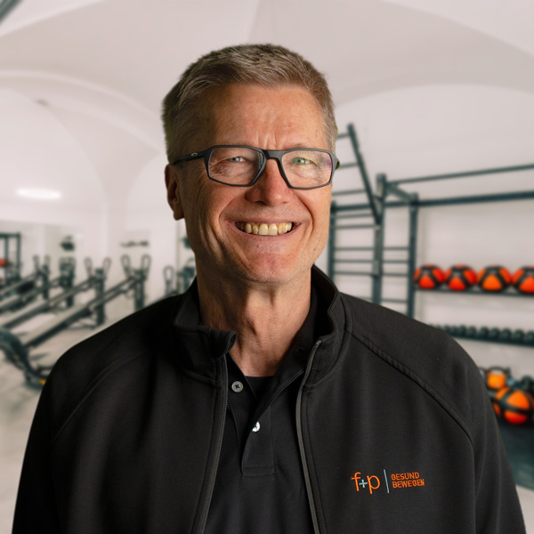 Martin Kr., leitender Arzt in Memmingen, trägt schwarze Jacke mit fp Gesund Bewegen Logo, Brille und lächelt vor einem Fitnessraum mit Hanteln.