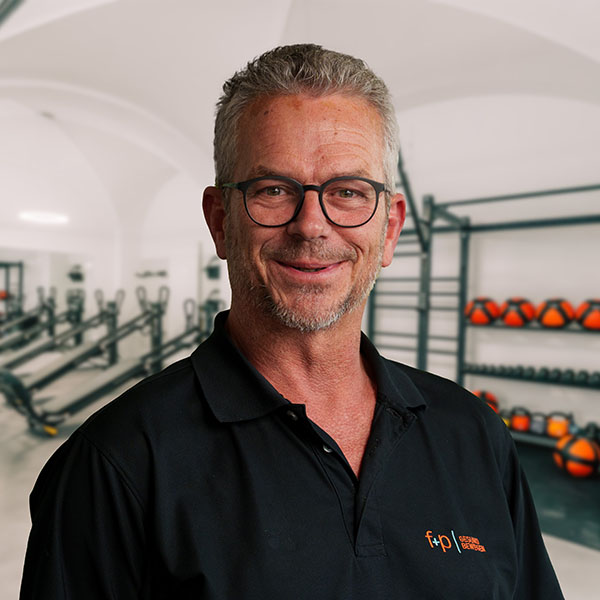 Mann mit Brille und schwarzem Polohemd steht in einem Fitnessstudio mit Trainingsgeräten im Hintergrund.