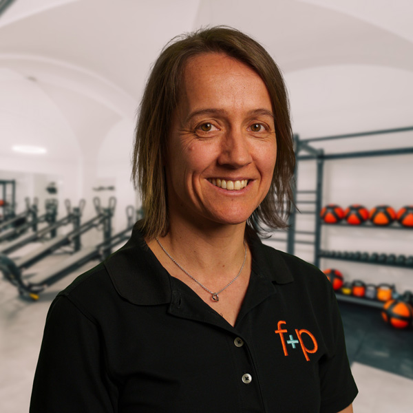 Martina Nebl Frau mit kurzem Haar in einem schwarzen Poloshirt lächelt vor einer Fitnessstudio-Hintergrund.