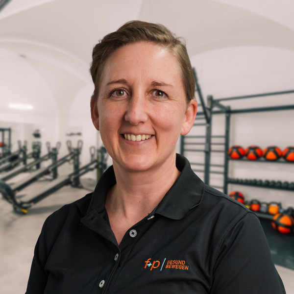 Martina in einem schwarzen Polohemd mit Logo von f+p Gesund Bewegen, Portrait im Trainingsraum des Allgäu-Standorts, Hintergrund mit Fitnessgeräten und roten Medizinbällen