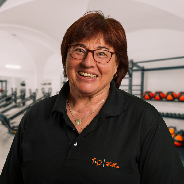 Monika Ra., Inhaberin von f+p Gesund Bewegen, lächelt in einem Trainingsraum. Sie trägt ein schwarzes Polo-Shirt mit dem Praxislogo, im Hintergrund Fitnessgeräte.