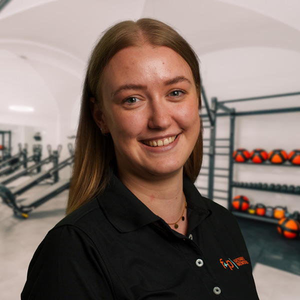 Nadine Holzer Eine Person mit langen, blonden Haaren trägt ein schwarzes Polohemd in einem Fitnessstudio.