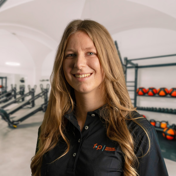 Nina von f+p Gesund Bewegen im Allgäu, blondes langes Haar, schwarze Polojacke mit Logo, Portrait im Trainingsstudio mit Trainingsgeräten im Hintergrund