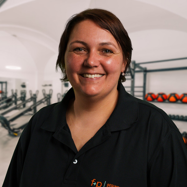 Sabrina Hu, lächelnde Mitarbeiterin von f+p Gesund Bewegen, trägt ein schwarzes Polo und steht in einem hellen Fitnessstudio mit Trainingsgeräten im Hintergrund.