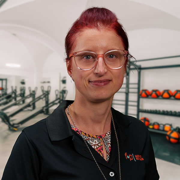 Eine Person mit rotem Haar und Brille, die ein schwarzes Polohemd trägt, steht in einem Fitnessstudio.