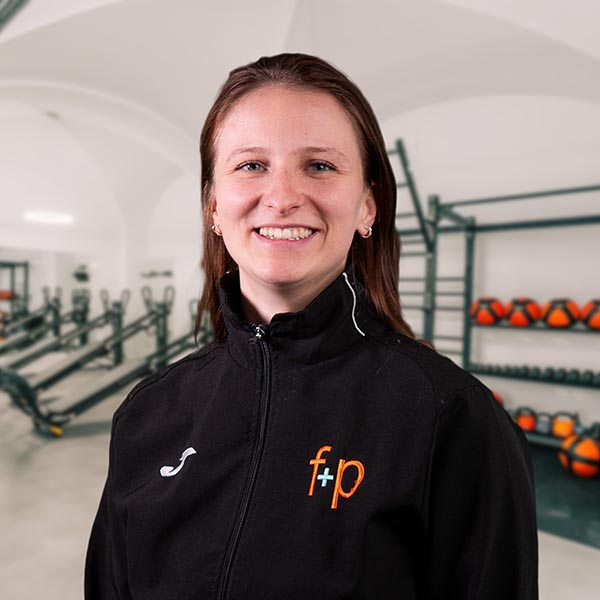 Frau mit langen Haaren in schwarzer Jacke mit Logo in einem Fitnessstudio.