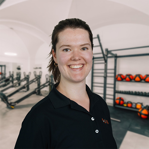 Im Physiobereich von f+p Gesund Bewegen im Allgäu lächelt Susanne in schwarzem Poloshirt vor Trainingsgeräten der Physiotherapie-Praxis.