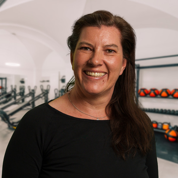 Susi Cossaro Frau mit langen, dunklen Haaren, die in einem Fitnessstudio vor einer Wand mit Gewichten steht.