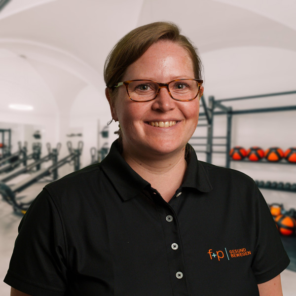 Tanja Da, eine lächelnde Physiotherapeutin mit Brille in schwarzer Polo-Kleidung vor Trainingsgeräten, Allgäu, f+p Gesund Bewegen, wirkt freundlich und professionell