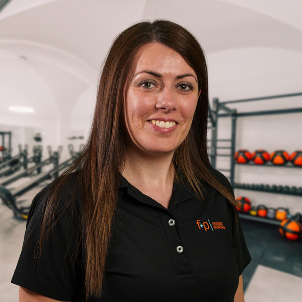 Teresa-Na, Teresa, im schwarzen Polo bei f+p Gesund Bewegen im Allgäu, lächelt in einem hellen Trainingsraum mit Fitnessgeräten und Reha-Ausstattung.