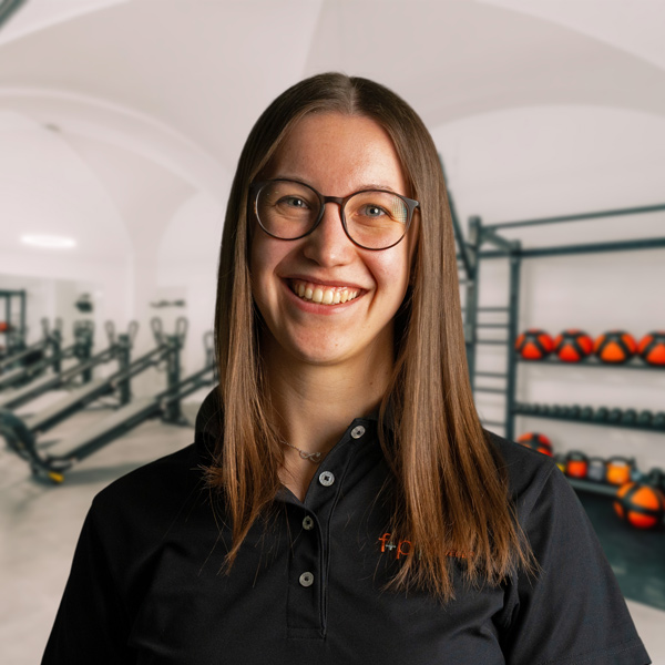 Theresa von f+p Gesund Bewegen im Allgäu mit Brille, lachendes Porträt vor Trainingsraum, Logo f+p auf schwarzer Bluse, Hintergrund mit Geräten