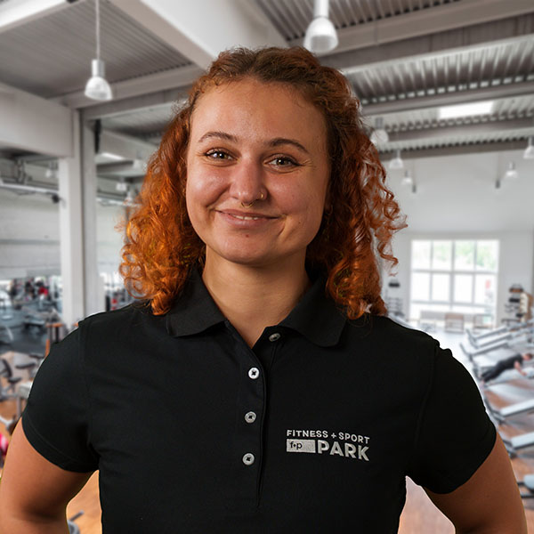 Junge Frau mit lockigem, orangefarbenem Haar in einem schwarzen Polohemd mit Fitness-Logo.