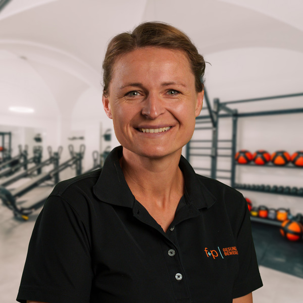 Veronika von f+p Gesund Bewegen im Allgäu, trägt ein schwarzes Polo-Shirt mit Logo und steht in einem hellen Fitnessraum mit Trainingsgeräten im Hintergrund, freundliches Lächeln
