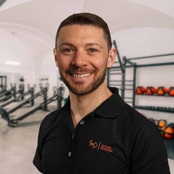 Walter Pe, Physiotherapeut bei f+p Gesund Bewegen, lächelt im Trainingsraum in einem schwarzen Polo; im Hintergrund befinden sich Fitnessgeräte und Medizinball-Reihen.