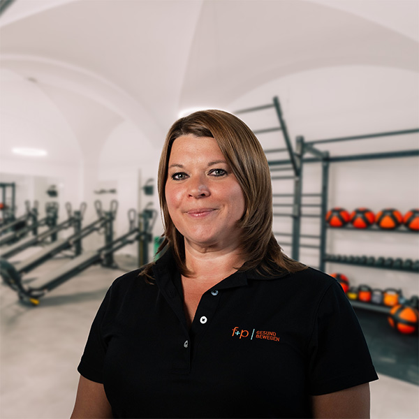 Frau mit braunen Haaren in schwarzem Poloshirt vor Fitnessgeräten in einem Studio.