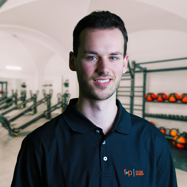 Adrian, Physiotherapeut bei f+p Gesund Bewegen, trägt dunkles, dunkellblaues Poloshirt; Hintergrund zeigt einen Trainingsraum mit Hanteln und Trainingsgeräten.