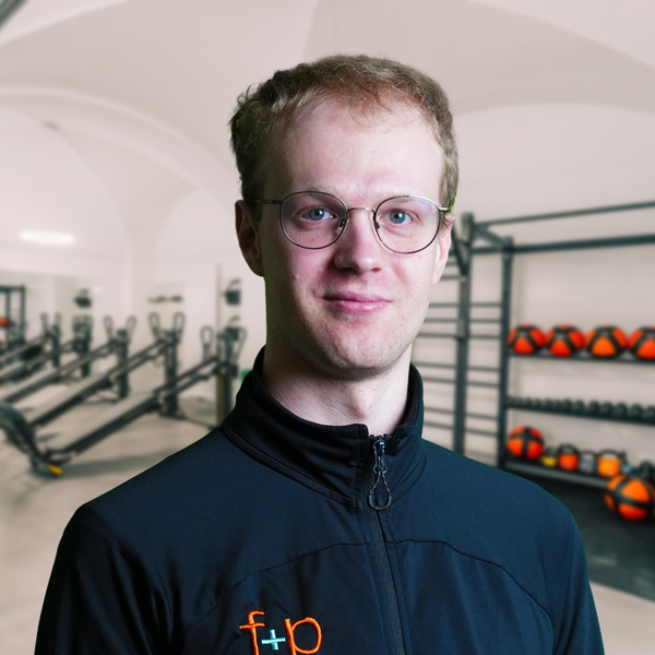 Adrian, Physiotherapeut bei f+p Gesund Bewegen, im dunkelblauen Trainingsjacke und Brille vor einem Trainingsraum mit Rudern und Medizinbällen.