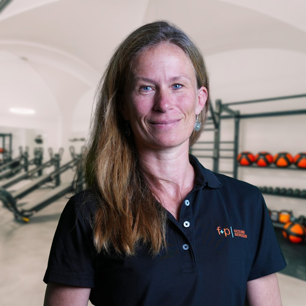 Frau mit langen Haaren in einem schwarzen Poloshirt steht in einem Fitnessstudio mit Trainingsgeräten im Hintergrund.