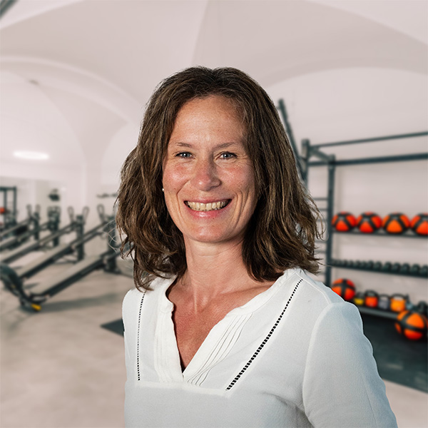 Frau mit lockigem Haar in einem Fitnessstudio, lächelnd vor einer Trainingsfläche.