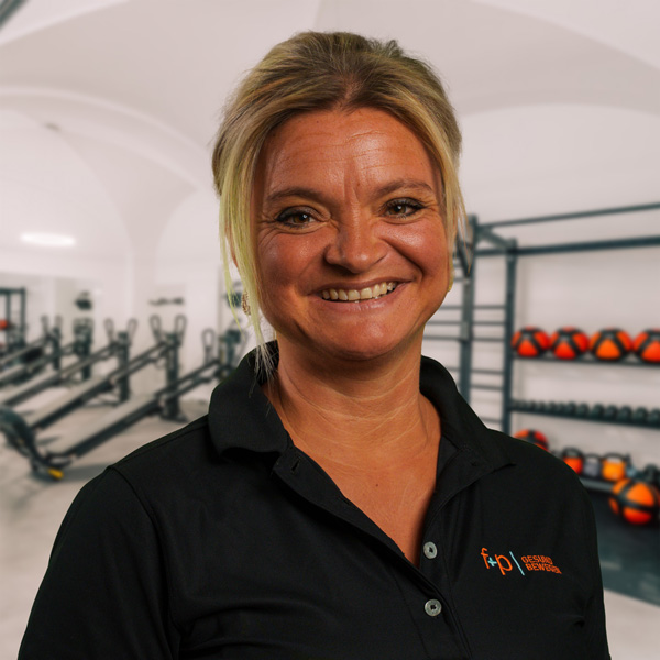 Carina, blondes Haar, lächelnd in schwarzer f+p Gesundheitsbewegen Polo, Hintergrund Fitnessstudio mit Geräten und Gewichten im Allgäu