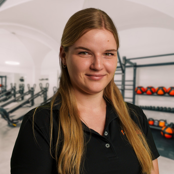 Carolin Le – Porträt einer jungen Frau mit hellbraunen Haaren in schwarzem Polo vor einem hellen Fitnessraum mit Trainingsgeräten im Allgäu