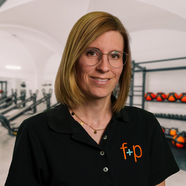 Corinna, Mitarbeiterin von f+p Gesund Bewegen, mit blondem Haar, Brille und schwarzem Polo, im Fitnessstudio vor Trainingsgeräten und Hanteln im Hintergrund.