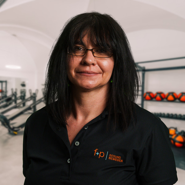 Daniela Ge, dunkelhaarige Mitarbeiterin von f+p Gesund Bewegen, trägt schwarze Bluse und Brille; im Hintergrund Trainingsgeräte und Hanteln in einem Studio