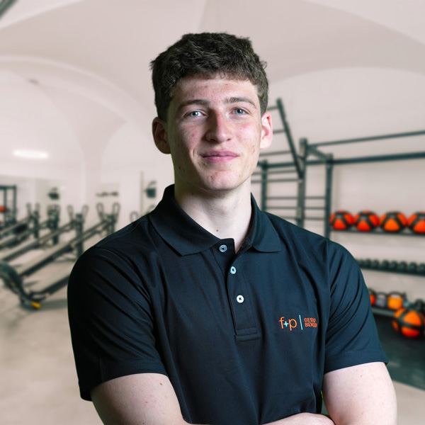 Emilio – junger Mann in schwarzem Polo-Shirt mit f+p-Logo steht vor einer Trainings- und Therapierumgebung in einer Allgäu-Praxis, klarer Fokus auf dem Gesicht und professioneller Ausstrahlung.