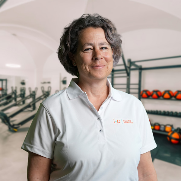 Hanna, Mitarbeitende bei f+p Gesund Bewegen, mit grauen Haaren in einem weißen Polo-Shirt mit dem Logo vor einer Trainingslandschaft und Fitnessgeräten im Allgäu-Haus