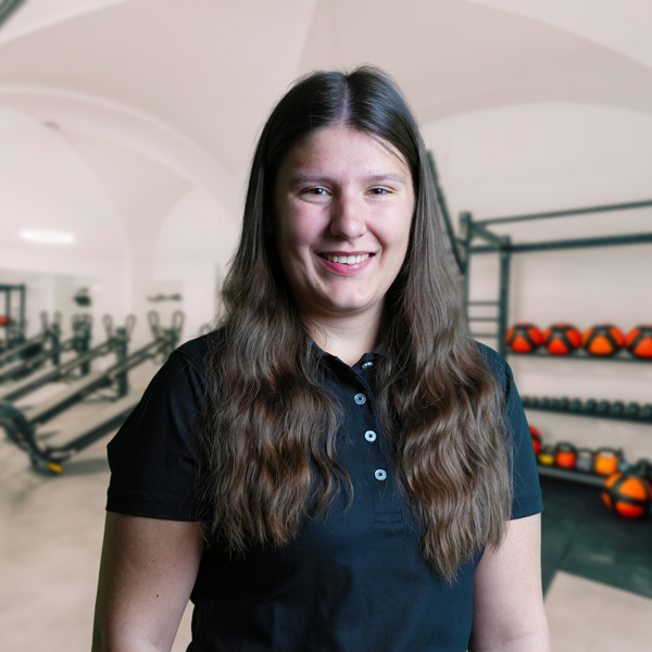 Eine Frau mit langen, braunen Haaren in einem Fitnessstudio, trägt ein schwarzes Polohemd.