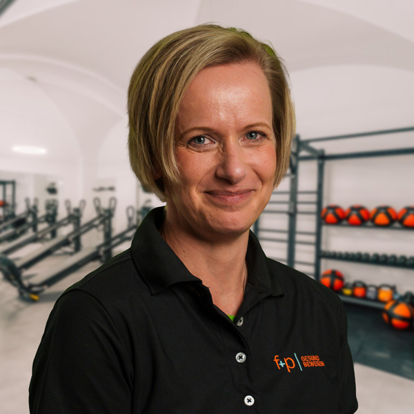 Kerstin, Mitarbeiterin von f+p Gesund Bewegen im Allgäu, mit kurzen blonden Haaren und schwarzem Polo vor einem Trainingsraum mit Hanteln und Medizinbällen, freundliches Lächeln