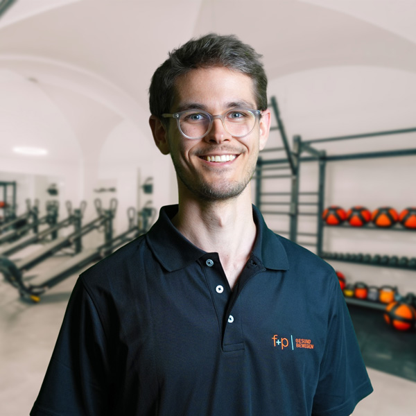 Lennart, Mitarbeiter von f+p Gesund Bewegen, steht lächelnd in einer Physiotherapie-Praxis im Allgäu. Er trägt ein dunkles Polohemd mit dem f+p Logo, wirkt freundlich und professionell, Brille auf der Nase.