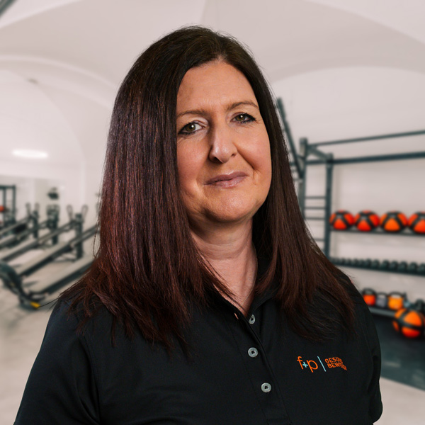 Marion Pröbstl Porträt einer Frau mit langen, braunen Haaren, die ein schwarzes Polohemd trägt und in einem Fitnessstudio steht.