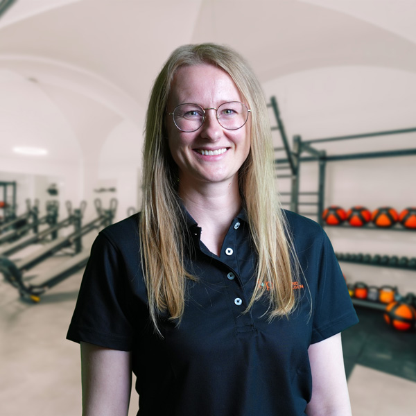 Monika Super Frau mit langen blonden Haaren und Brille in einem schwarzen Poloshirt lächelt in einem Fitnessraum.
