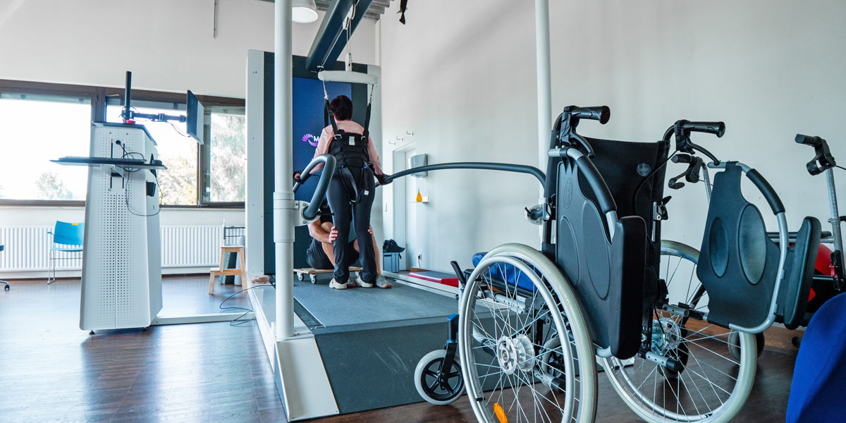 Rehabilitationsgerät mit einem Patienten, der Unterstützung beim Stehen erhält, sowie einem Rollstuhl im Raum.