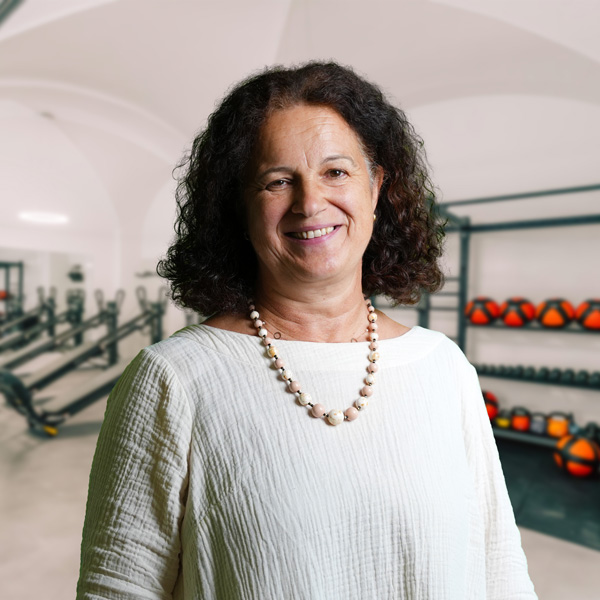 Silvia, eine Fachärztin für psychotherapeutische Medizin, mit dunklen Locken, weißem Oberteil und Perlenkette, vor Trainingsgeräten im Allgäu-Fitnesszentrum von f+p Gesund Bewegen