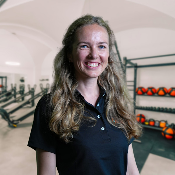 Frau mit langen, lockigen Haaren in einem schwarzen Polohemd lächelt vor einer Fitnessumgebung.