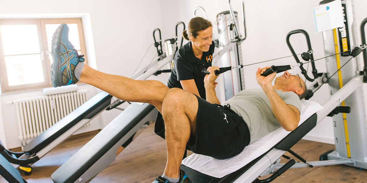Ein Mann trainiert an einem Fitnessgerät mit Unterstützung eines Trainers in einem Trainingsraum.