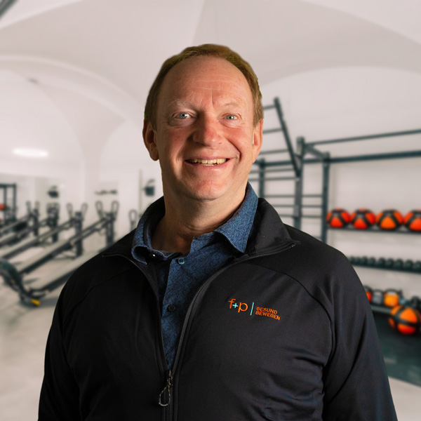 Mann in schwarzer Sportjacke lächelt vor einer Fitnessumgebung mit Geräten und Gewichten.