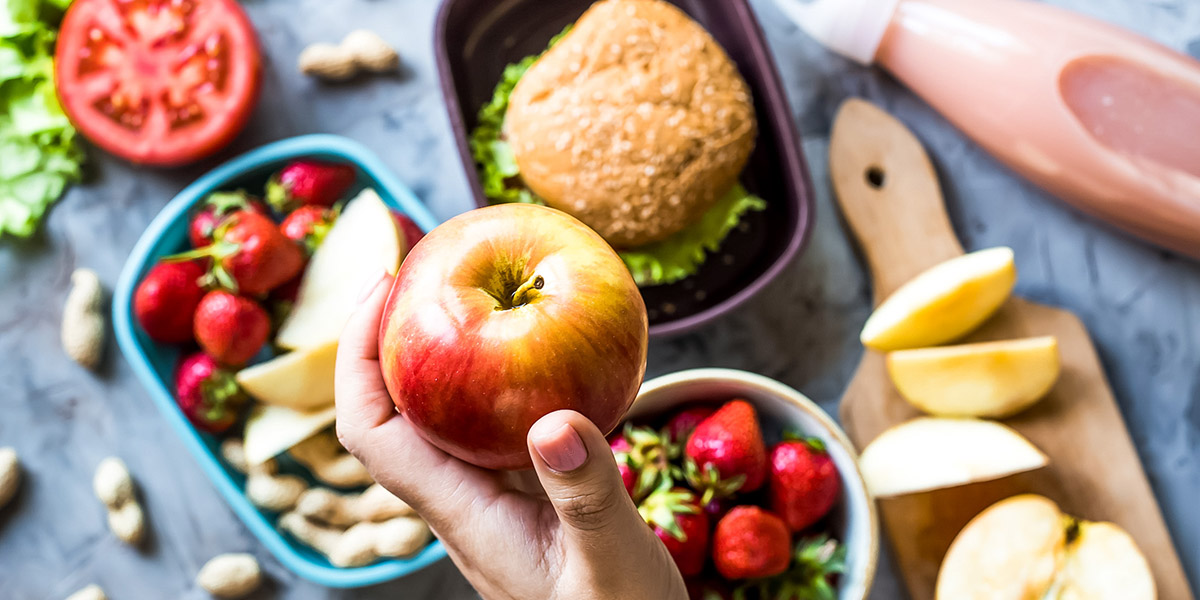 Eine Hand hält einen roten Apfel über einer Auswahl an Lebensmitteln, darunter ein Burger und Obst.