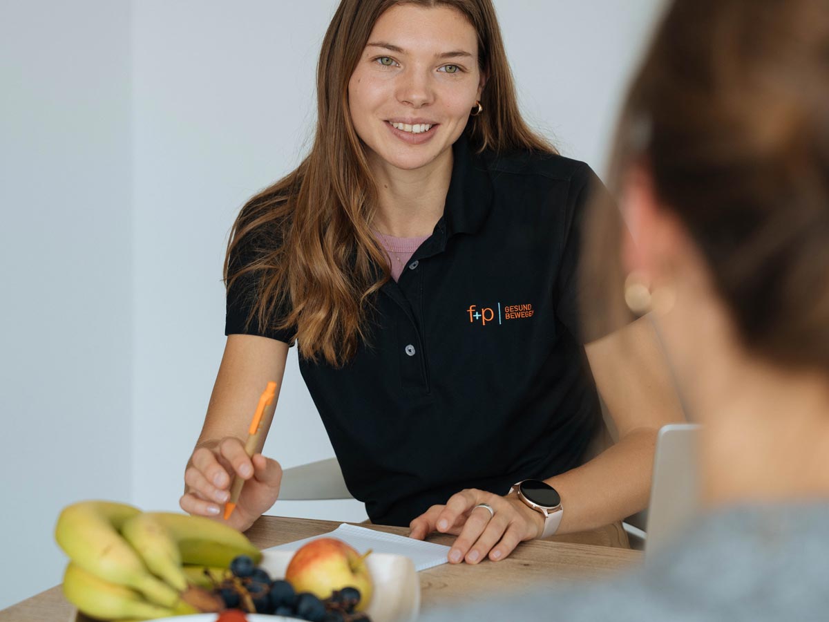 FUP00199_web Junge Frau in schwarzem Poloshirt mit f+p Logo, lächelnd und Notizen machend, während sie mit einer anderen Person spricht.