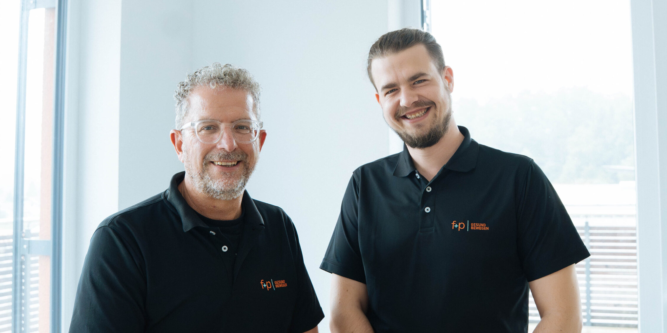 Zwei Männer in schwarzen Poloshirts mit dem Logo von f+p Gesund Bewegen stehen nebeneinander und lächeln.
