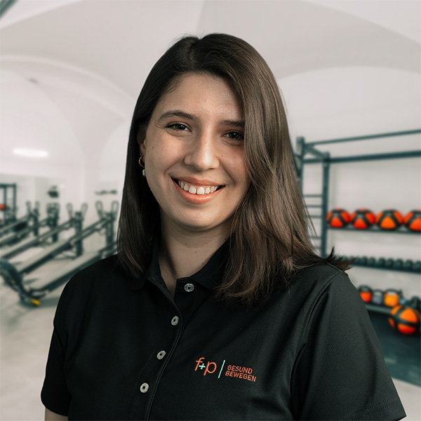 Gizem, Mitarbeiterin von f+p Gesund Bewegen, lächelt im Fitnessstudio. Sie trägt ein schwarzes Polo-Shirt mit dem Logo und hat braune Haare.