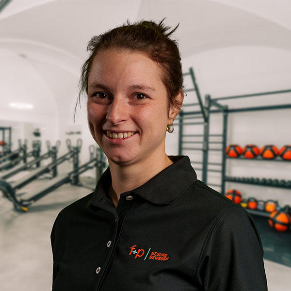Iris, Mitarbeiterin von f+p Gesund Bewegen, in schwarzem Polohemd, Portrait im Trainingsraum mit Kraftgeräten, Hanteln und Bindegliedern im Hintergrund
