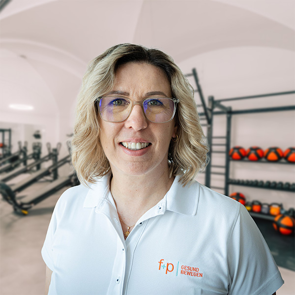 Blonde Mitarbeiterin Marta mit Brille im weißen Polo von f+p Gesund Bewegen vor Trainingsbereich, freundlicher Gesichtsausdruck und klarer Blick.